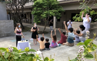 children's ministry director Cayanna Herrera leads children in a song with her ukulele. Children's backs are to the camera and there arms are raised, moving to the song.