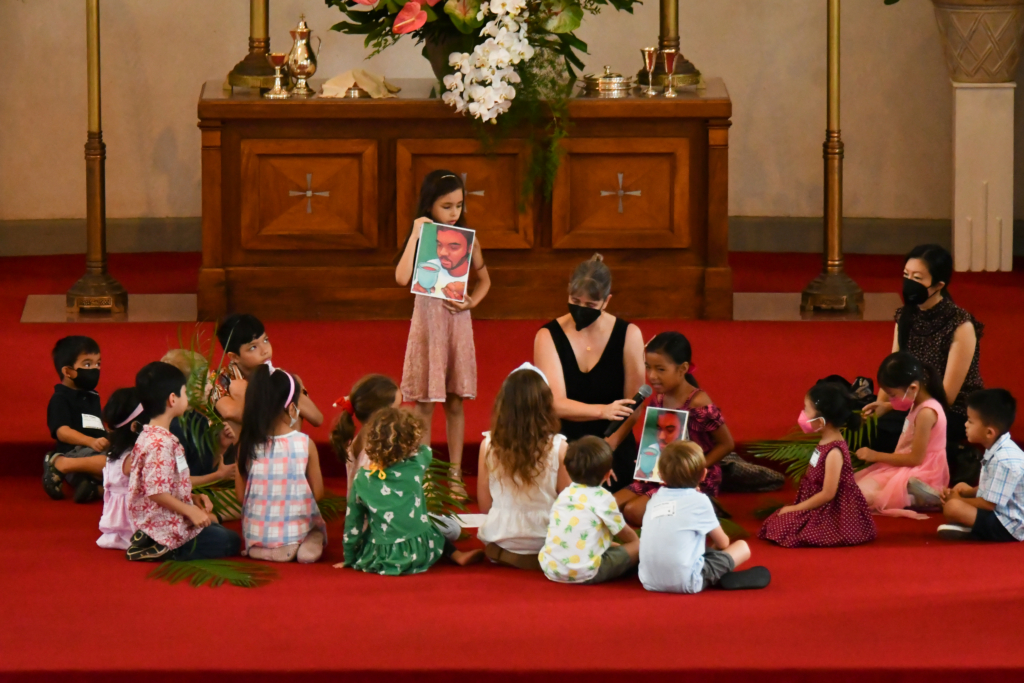 children sitting on sanctuary chancel gathered around for time with keiki during worship service two young girls hold up paintings of jesus