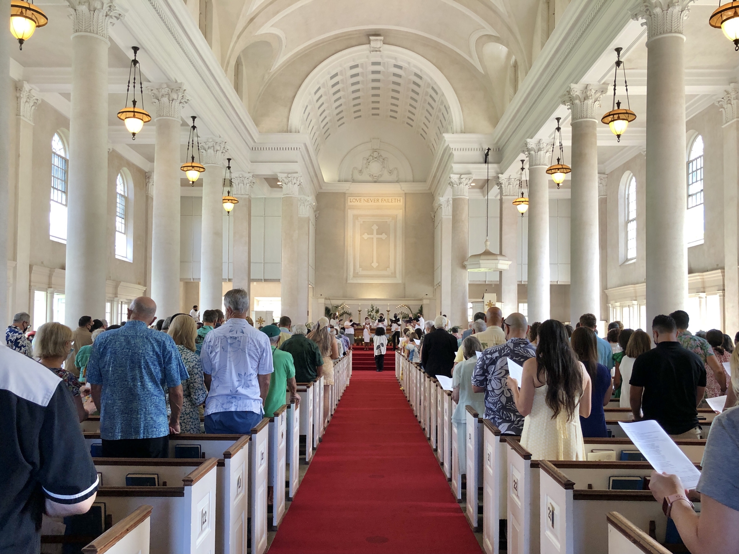 people standing in pews in sanctuary taken from middle of aisle looking forward facing people's backs